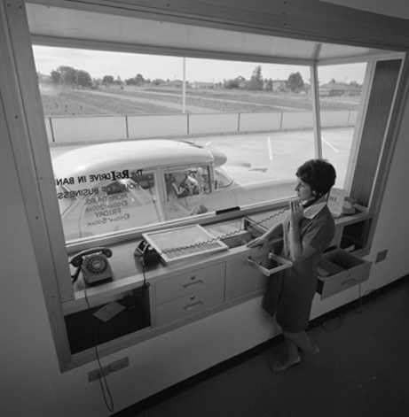black and white photo of a drive in bank view from inside the bank where a staff member stands at the glass ready to serve the car on the other side