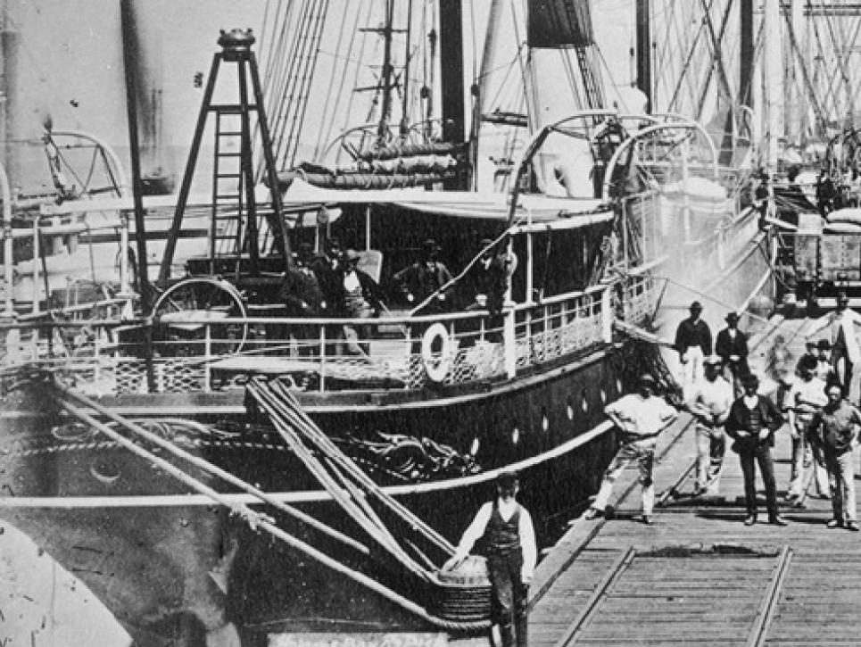 Black and white photo of people on pier with tall ships and pier donkey.